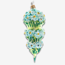 A vintage-style glass Christmas ornament shaped like a tiered cupcake, featuring light blue and green frosting, white daisy flowers, and small gold accents, shown against a plain light gray background.