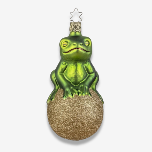 A green frog Christmas ornament sitting on a glittery gold egg, topped with a small gold crown and star, shown against a light gray background.