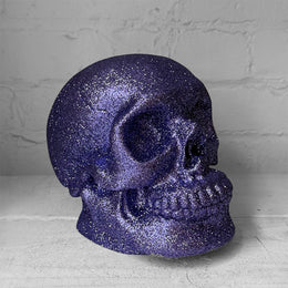 A glittery purple skull sculpture with a textured, sparkly finish, displayed against a light gray brick wall background.