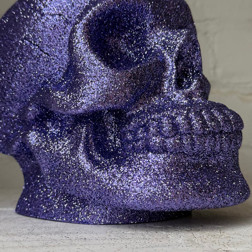 A close-up of a glittery purple skull decoration, showing its textured, sparkly surface against a neutral light gray background.