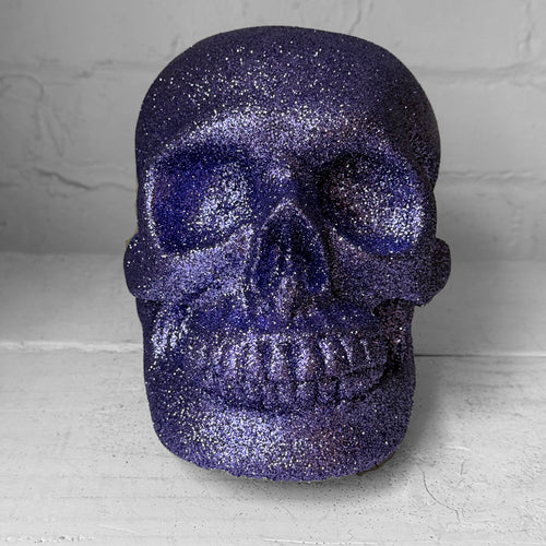 A glittery skull decoration in a deep purple color, set against a light gray brick wall background.