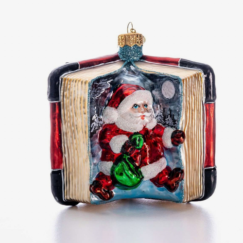 A hand-painted glass Christmas ornament shaped like an open book, featuring Santa Claus sitting inside and holding a green sack. The ornament has a gold hanging loop at the top and is photographed on a clean white background.