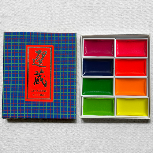 A boxed set of eight Chinese watercolor palettes shown on a light fabric background, with the lid on the left and the open tray of colorful watercolor blocks on the right.