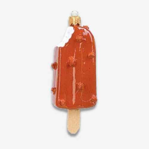 A novelty Christmas ornament shaped like a popsicle, featuring a glossy orange-red body with white frosting on the top half, small raised red dots, and a light wood-look stick, shown on a plain light gray background.