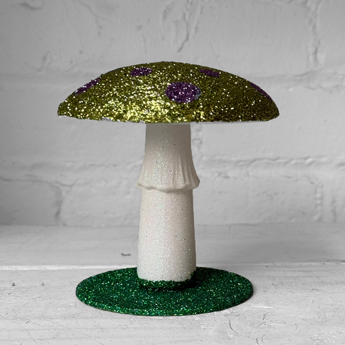 A decorative mushroom figurine with a glittery olive green cap, purple glitter spots, a white glittery stem, and a round green glitter base, set against a white brick wall background.