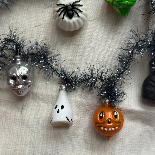 A close-up of a Halloween ornament garland featuring a silver skull, a white ghost, an orange jack-o'-lantern, and a black spider decoration, draped on black tinsel against a beige fabric background.