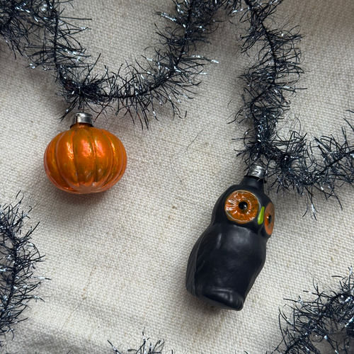 Two Halloween-themed glass ornaments on a beige fabric background with black tinsel. One ornament is a glossy orange pumpkin, and the other is a black owl with large orange and green eyes.