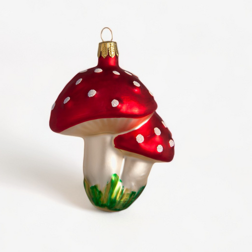 A decorative glass Christmas ornament shaped like two red mushrooms with white spots and green grass at the base, shown on a plain white background.