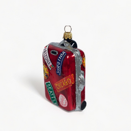 A miniature red suitcase Christmas ornament decorated with travel stickers and a gold hanging loop, shown on a clean white background.